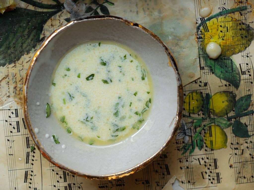 A bowl of vegan "egg wash" with chopped herbs, placed on a vintage music sheet with lemon illustrations. A few splatters of soup are visible around the bowl.